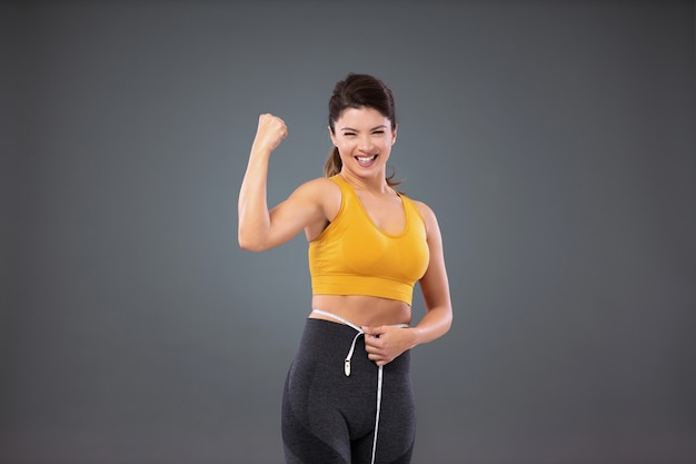 Uma mulher em forma atlética em roupas esportivas em frente a uma parede cinza medindo sua cintura com uma fita adesiva