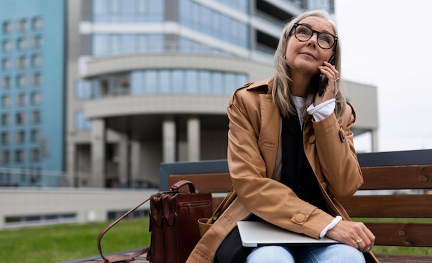 Uma mulher em idade de aposentadoria com um laptop fala em um celular perto do escritório