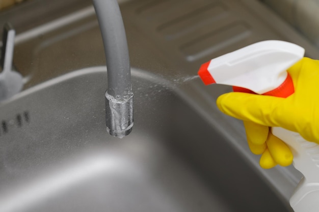 Uma mulher em luvas de borracha limpa a torneira e pia na cozinha aplicando desinfetante de um frasco de spray Closeup