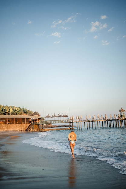 Uma mulher em um maiô e um chapéu com óculos caminha ao longo da praia ao pôr do sol. O conceito de recreação marítima. Foco seletivo
