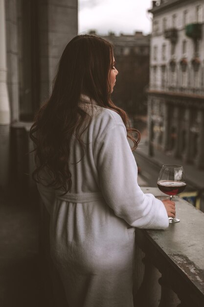 Uma mulher em um roupão branco com uma taça de vinho em pé de costas