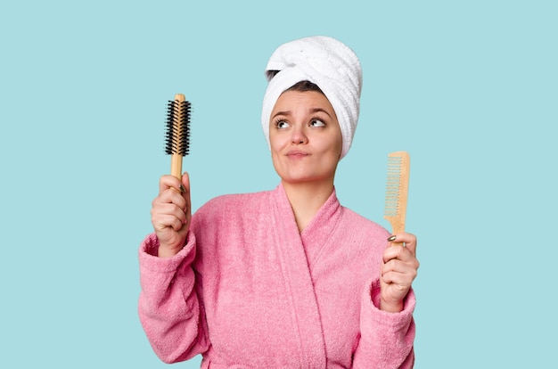 Uma mulher em um roupão rosa segurando uma escova de cabelo e um pente com uma toalha enrolada na cabeça