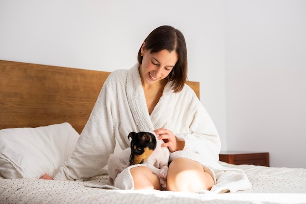 Uma mulher em um roupão senta-se em uma cama com seu cachorro