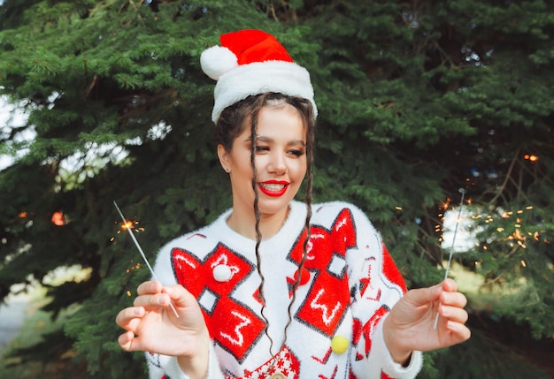 Uma mulher em um suéter de inverno com um chapéu de Papai Noel e lábios vermelhos segura um diamante contra o fundo das árvores de Natal ano novo natal