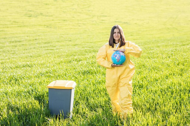 Uma mulher em um traje de proteção amarelo fica no meio de um campo verde e segura um globo nas mãos ao lado de uma lata de lixo