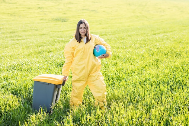 Uma mulher em um traje de proteção amarelo fica no meio de um campo verde segurando um globo e com a outra mão carrega uma lata de lixo O conceito de salvar o planeta da poluição ambiental