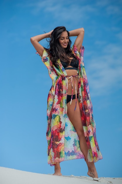 Uma mulher em um vestido colorido fica em uma praia em frente a um céu azul.