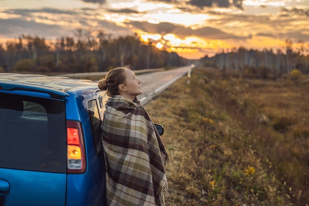 Uma mulher em uma manta fica ao lado do carro na beira da estrada no fundo do conceito de viagem ao amanhecer