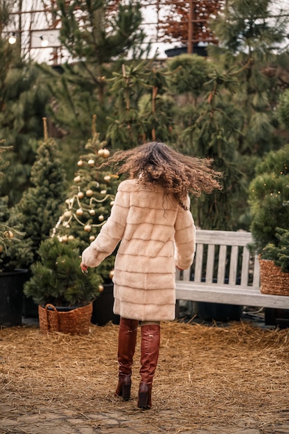 Uma mulher encaracolada sorridente com um casaco de pele está circulando entre as árvores de Natal na rua Esperando o Natal e o Ano Novo
