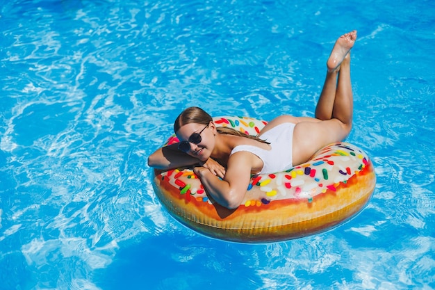 Uma mulher esbelta em um maiô branco e óculos de sol está na piscina em um círculo inflável Descanso de verão