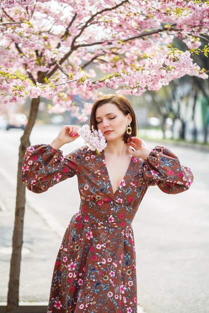 Uma mulher espetacular em um vestido brilhante está contra o pano de fundo de sakura. Uma mulher de cabelos escuros em uma linda roupa sorri na rua enquanto caminha