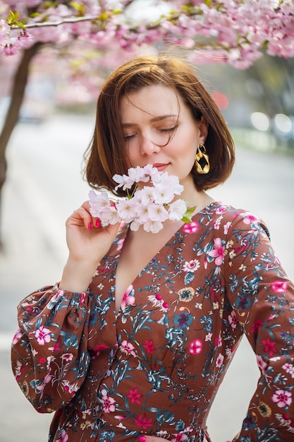 Uma mulher espetacular em um vestido brilhante está contra o pano de fundo de sakura. Uma mulher de cabelos escuros em uma linda roupa sorri na rua enquanto caminha