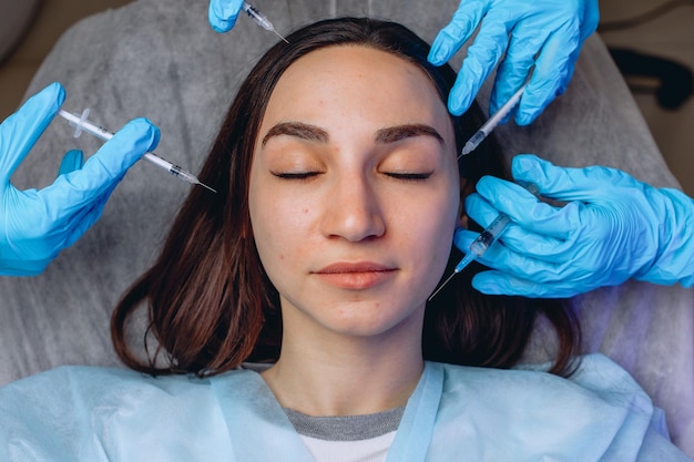 Uma mulher está deitada em um sofá branco. As mãos do cosmetologista enluvado seguram seringas e a cercam para o procedimento.