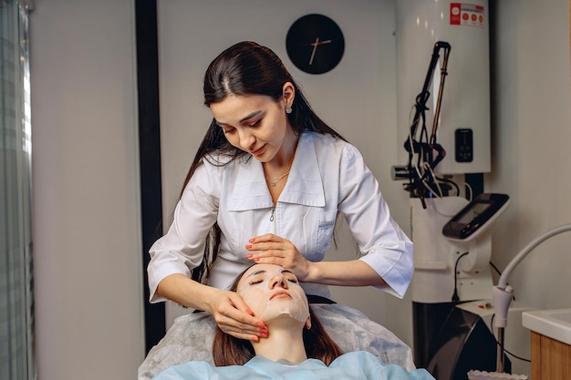 Uma mulher está deitada em uma cadeira em um salão de beleza com uma máscara facial hidratante e calmante enquanto um cosmetologista faz uma massagem.