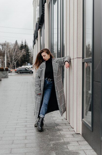 Uma mulher está parada na calçada em frente a uma casa com um casaco cinza