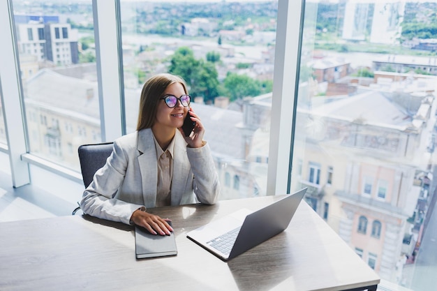 Uma mulher europeia alegre loura de óculos em roupas casuais elegantes está sentada em uma mesa com um laptop fazendo papelada e falando ao telefone Mulher de negócios no local de trabalho no escritório