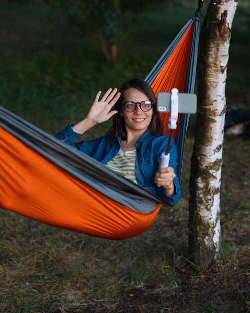 Uma mulher europeia em uma rede na natureza fala via link de vídeo com uma webcam uma blogueira de viagens