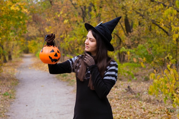 Uma mulher fantasiada de bruxa segura uma abóbora com doces na floresta de outono.