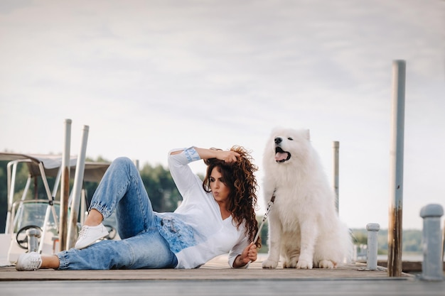 Uma mulher feliz com um grande cachorro branco encontra-se em um píer perto do mar ao pôr do sol.