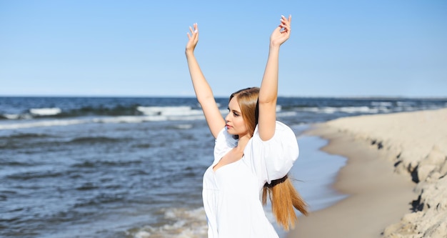 Uma mulher feliz e bonita na praia do oceano, de pé com um vestido branco de verão, a levantar as mãos.