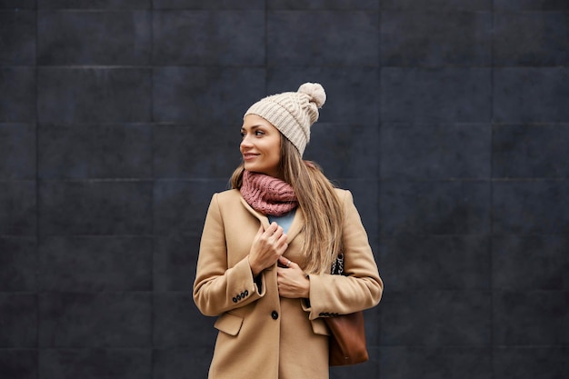 Uma mulher feliz e elegante em um cachecol e gorro de pé na rua e ajustando o casaco