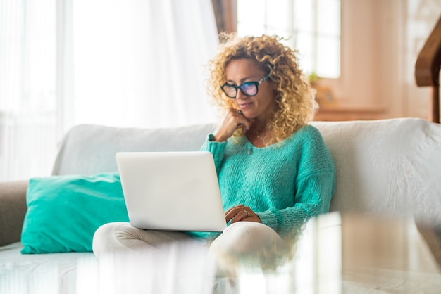 Uma mulher feliz e encaracolada sentada no sofá de sua casa usando o PC do computador navegando na internet - linda senhora usando um laptop no sofá de sua casa