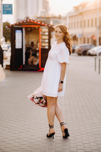 Uma mulher feliz em um vestido branco ao pôr do sol com um buquê de flores na cidade