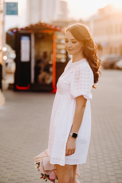 Uma mulher feliz em um vestido branco ao pôr do sol com um buquê de flores na cidade