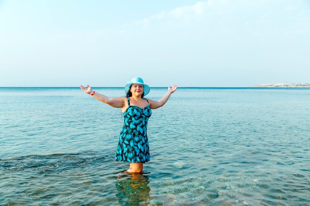 Uma mulher feliz em um vestido de verão azul e um chapéu está no mar com água até os joelhos.