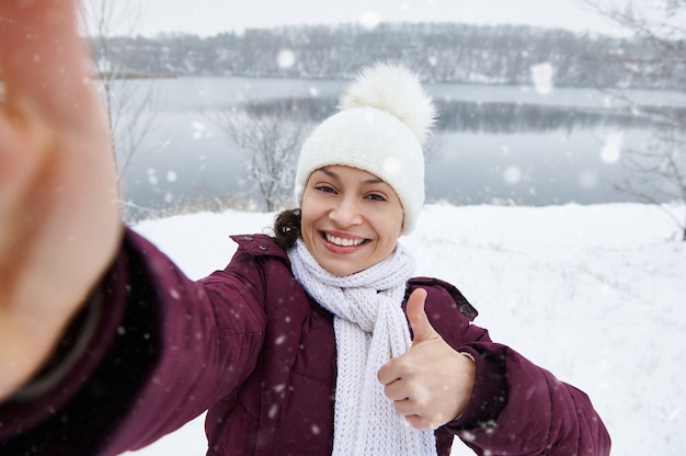 Uma mulher feliz mostrando o polegar e sorrindo para a câmera enquanto faz uma selfie curtindo uma queda de neve