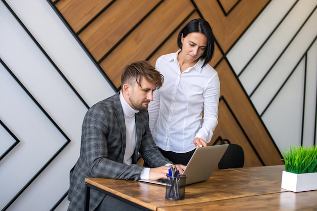 Uma mulher fica ao lado de um homem sentado em uma mesa com um laptop no escritório