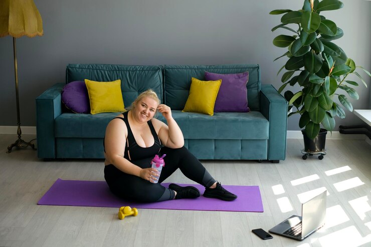 Foto uma mulher gorda feliz cansada está sentada em um tapete de treinamento segurando uma garrafa de água na mão