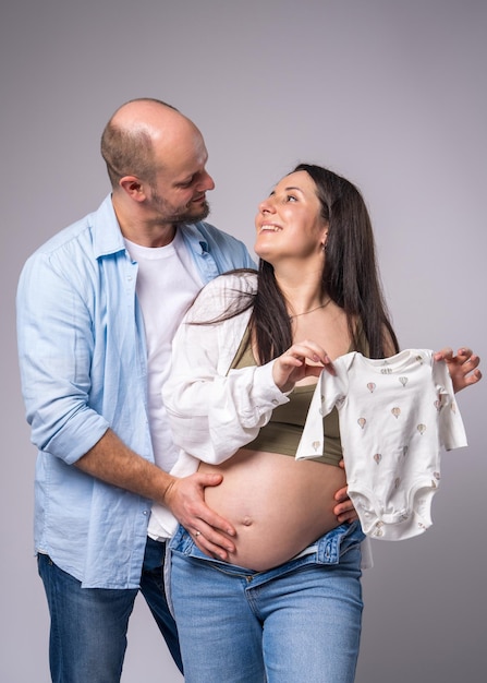 Uma mulher grávida com a barriga exposta ao lado de seu marido segurando roupas de bebê em suas mãos