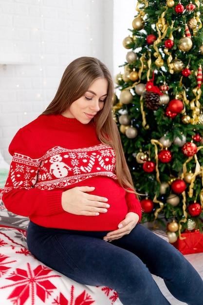 Uma mulher grávida com um suéter vermelho está acariciando sua grande barriga sob a árvore de Natal em casa e sonhando com um bebê e curtindo o ano novo e o Natal