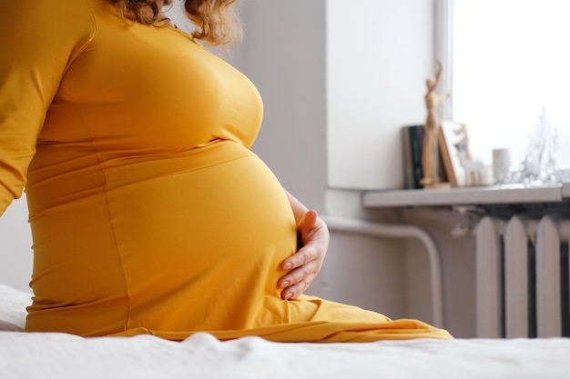 Uma mulher grávida com um vestido amarelo senta-se na cama e acaricia a barriga Fechamento da barriga