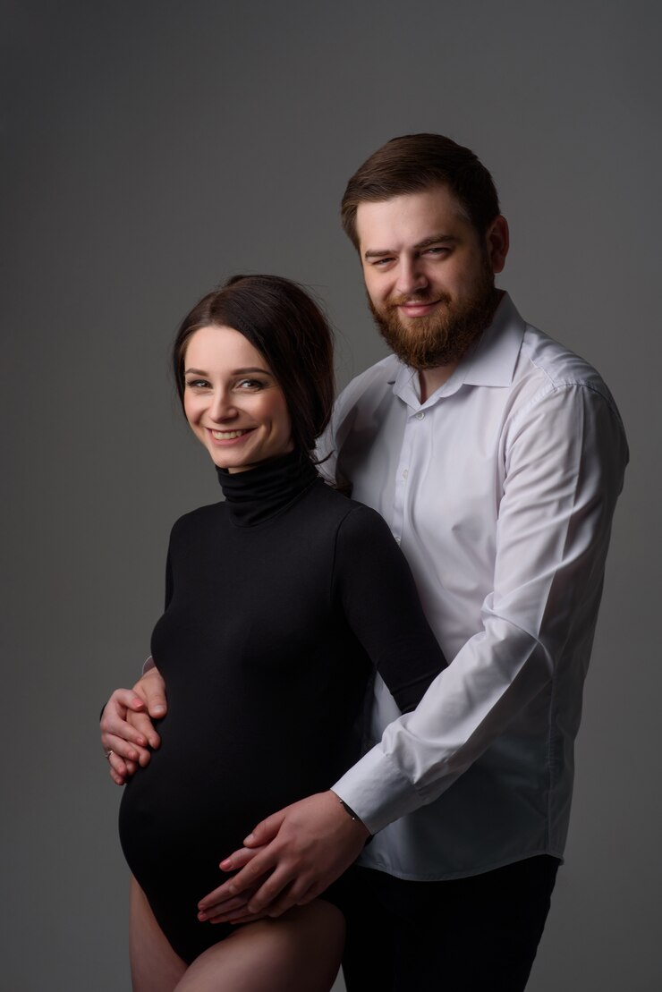Foto uma mulher grávida e seu marido estão abraçando em uma parede cinza casal esperando um bebê.