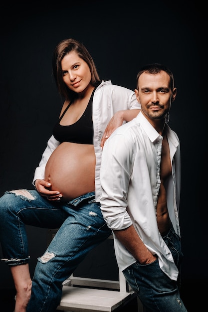 Uma mulher grávida e um homem em uma camisa branca e calça jeans em um estúdio em um fundo preto.