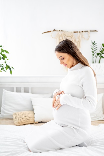 Uma mulher grávida em um vestido branco com uma barriga grande o acaricia com a mão A futura mãe está esperando e se preparando para o nascimento de uma criança em casa em uma cama em um quarto iluminado