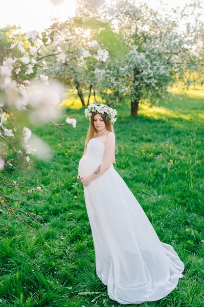 Uma mulher grávida em um vestido branco e uma guirlanda floral. Mulher grávida em um jardim botânico de florescência perto de sakura e macieiras