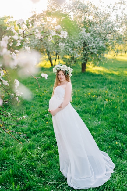 Uma mulher grávida em um vestido branco e uma guirlanda floral. mulher grávida em um jardim botânico de florescência perto de sakura e macieiras