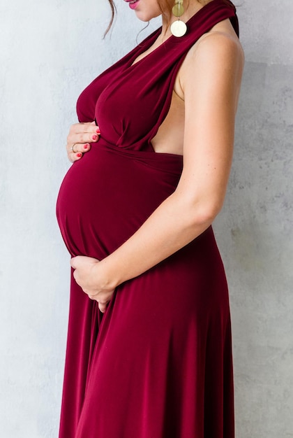 Uma mulher grávida em um vestido longo de seda cor de vinho abraça a barriga e posa em um fundo cinza