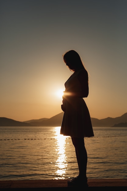 Uma mulher grávida está na praia ao pôr do sol e acaricia suavemente sua barriga, atrás dela estão