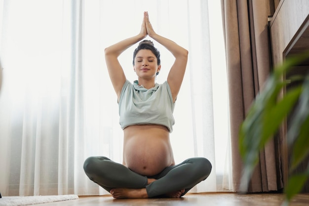 Uma mulher grávida feliz e calma está sentada no chão em casa e meditando A mulher grávida está fazendo ioga