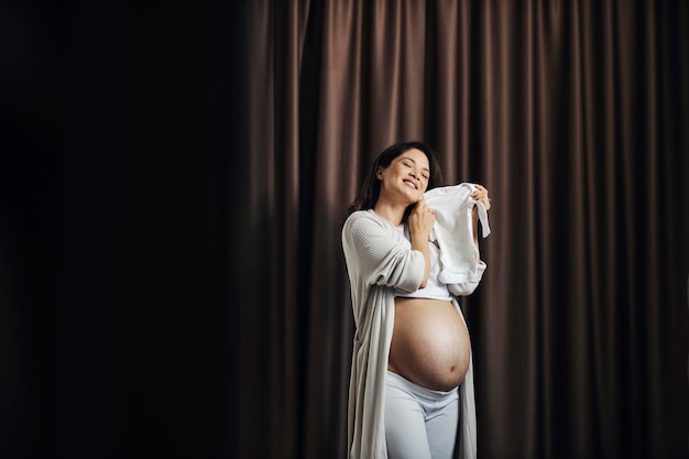 Uma mulher grávida feliz está de pé com a barriga nua com estrias e segurando o bebê onsie