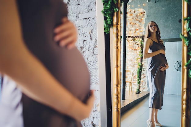 Foto uma mulher grávida feliz olha com amor para o reflexo dela mesma e de seu bebê no espelho.