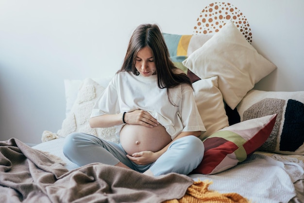 Uma mulher grávida sentada na cama em roupas domésticas segurando suavemente sua barriga grávida
