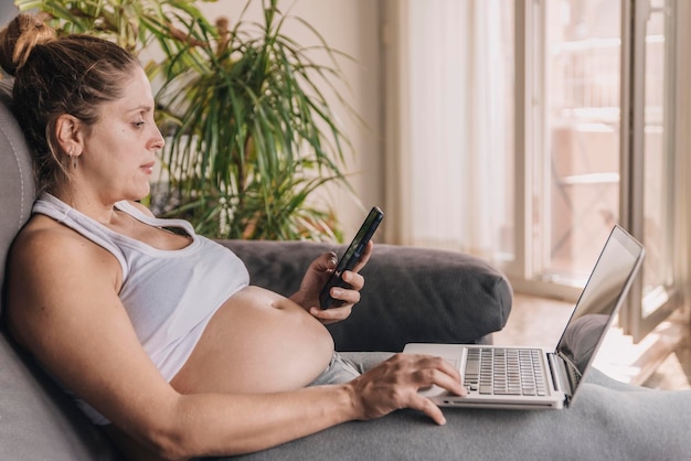 Foto uma mulher grávida sentada no sofá usando um laptop e smartphone