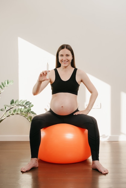 Uma mulher grávida sorridente está sentada em uma bola de exercícios e posando com uma seringa