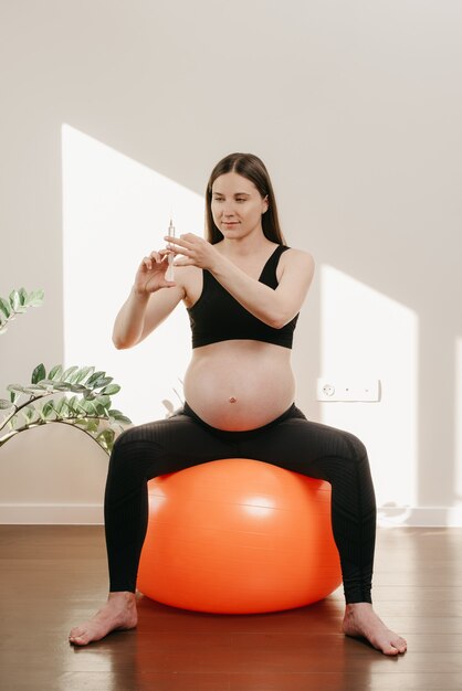 Uma mulher grávida sorridente está sentada em uma bola laranja de exercícios e verificando uma seringa com vitaminas em casa. Gravidez saudável.