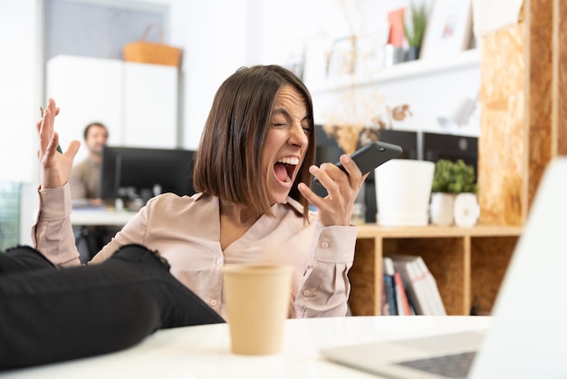 Uma mulher hispânica furiosa com os pés na mesa gritando ao telefone no escritório